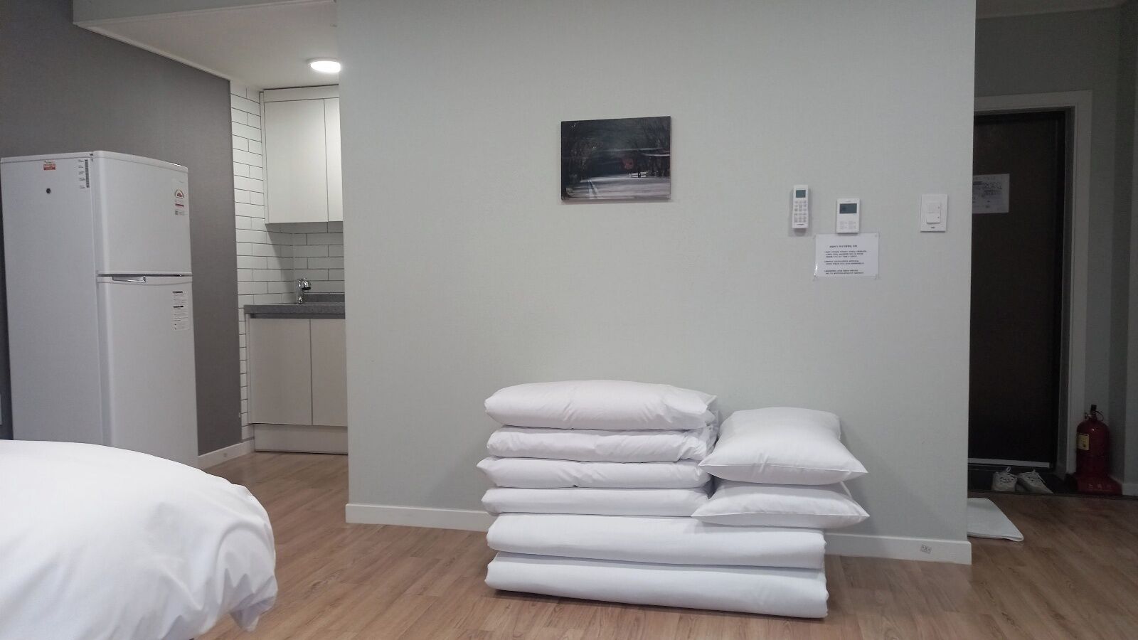 A hotel room with a bed, a stack of white pillows, a kitchen area, and a fire extinguisher.