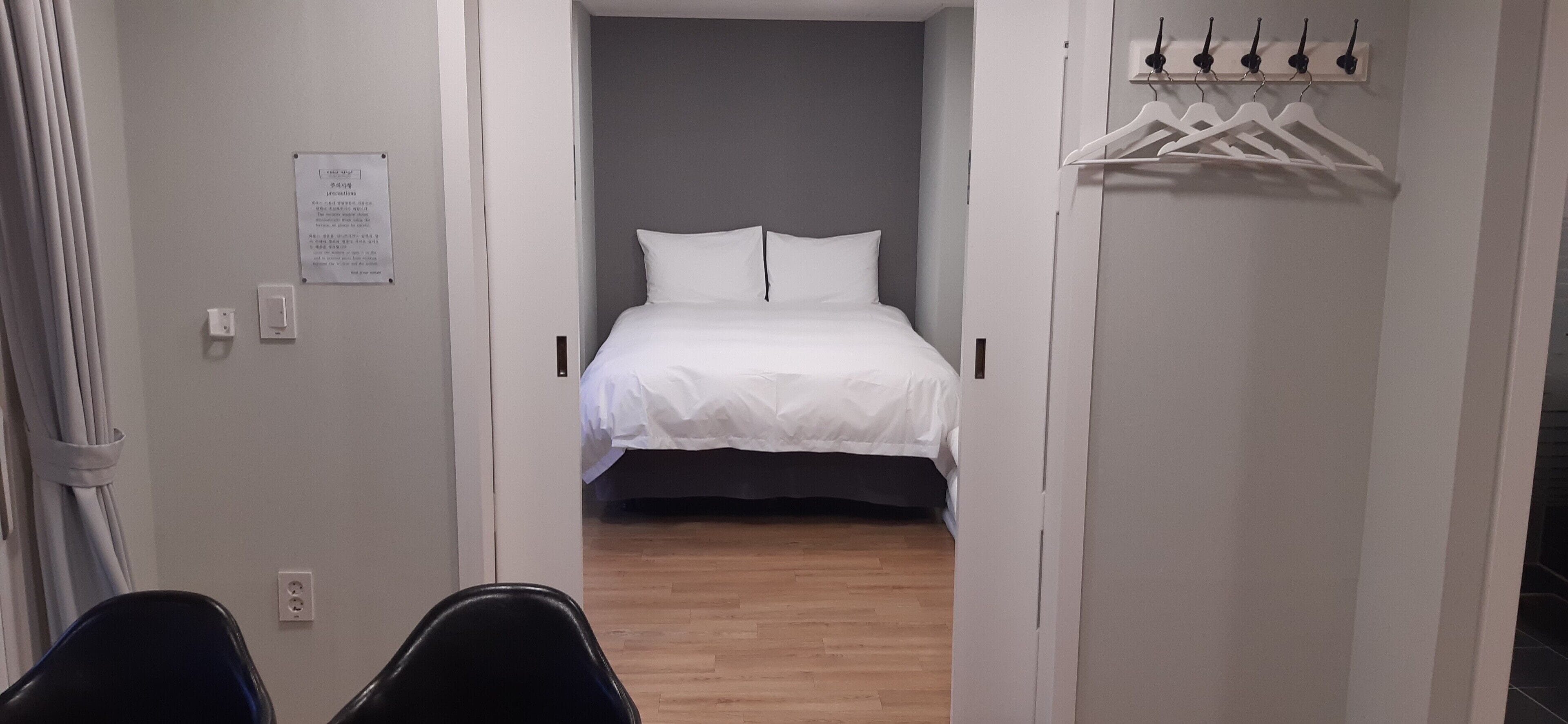 A hotel room with a bed, a coat rack with hangers, a door with a sign, and a chair.