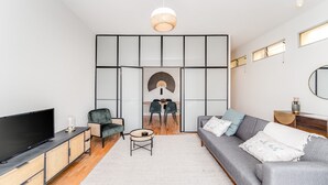 A modern living room with a flat-screen TV, a grey sofa, a round coffee table, and a dining area with chairs and a table. - Nomad's By Sta Catarina Flats - 3BDR Family (Porto)
