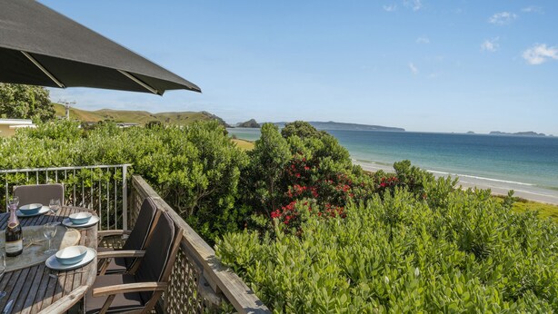 A wooden deck with a table set for dining, overlooking a beach and ocean. - Double the Beach - Opito Bay Holiday Home (Opito Bay)