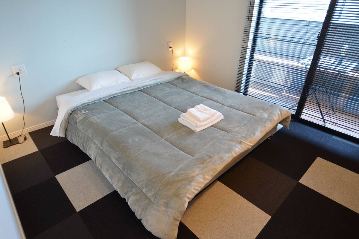 Ein ordentlich bezogenes Bett mit einer grauen Daunendecke, weißen Kissen und einem gefalteten Handtuch. Links befindet sich eine Nachttischlampe und rechts ein Fenster mit Jalousien.