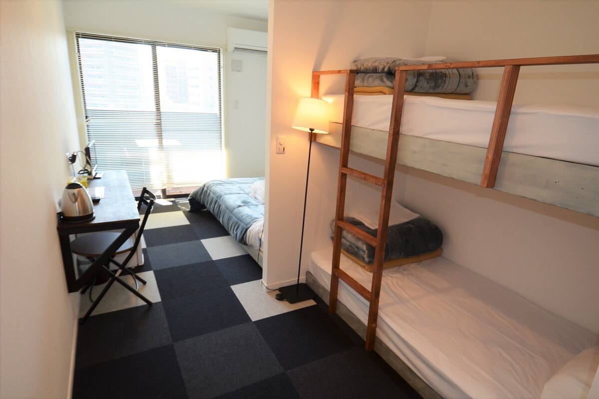 Ein kleiner, kompakter Raum mit einem Etagenbett, einem Schreibtisch mit Stuhl, einer Lampe und einem Fenster mit Jalousien.