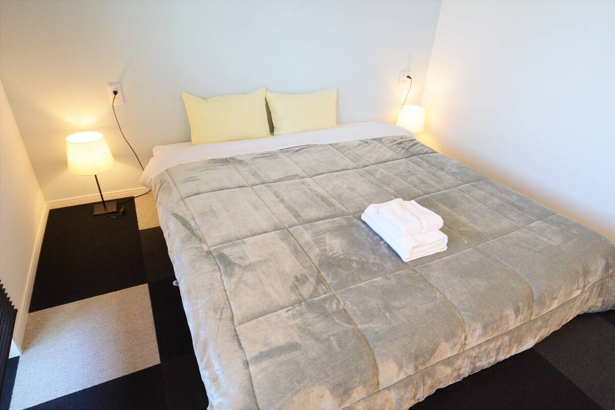 Ein ordentlich bezogenes Bett mit einer grauen Bettdecke, zwei Kissen und einem zusammengelegten weißen Handtuch. Ein Nachttisch mit einer Lampe steht daneben, und ein schwarz-weiß gestreifter Teppich liegt auf dem Boden.