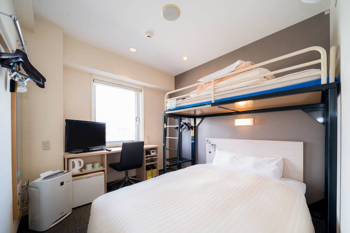 Une chambre d’hôtel avec un lit superposé, un bureau avec un ordinateur et une télévision, une chaise, un petit radiateur et une fenêtre.