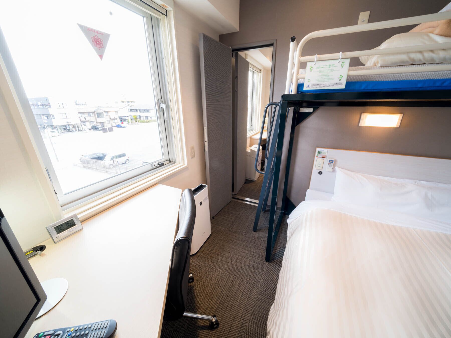 Quarto de hotel com cama beliche, uma escrivaninha com cadeira, janela com vista para a rua e mesa de cabeceira com controle remoto.