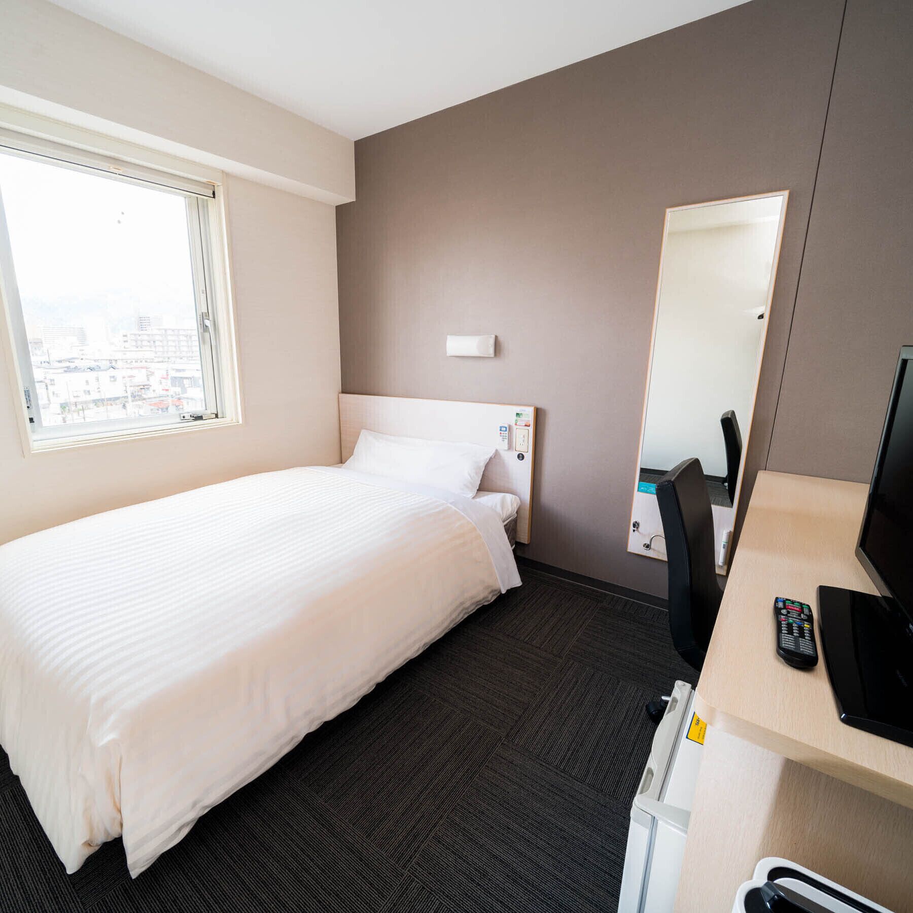 A hotel room with a bed, a desk with a computer and a TV, a chair, and a window with a city view.
