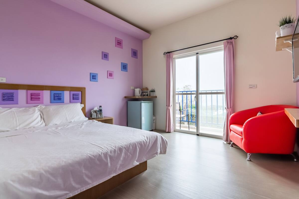 A hotel room with a bed, a red chair, a small table, a window with curtains, and a balcony with a view.