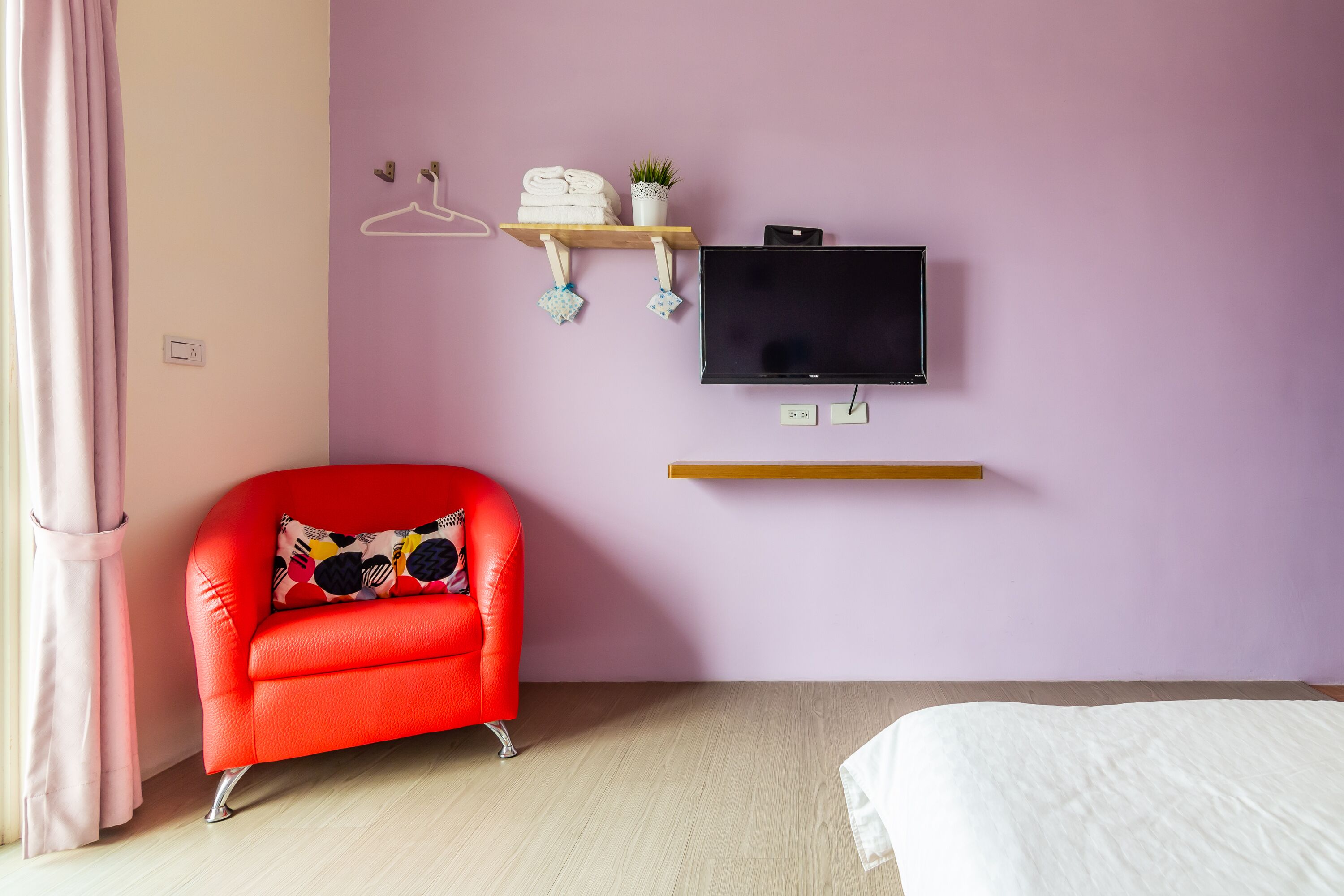 A room with a red armchair, a bed, a TV, a shelf with towels and a plant, and a pink wall.