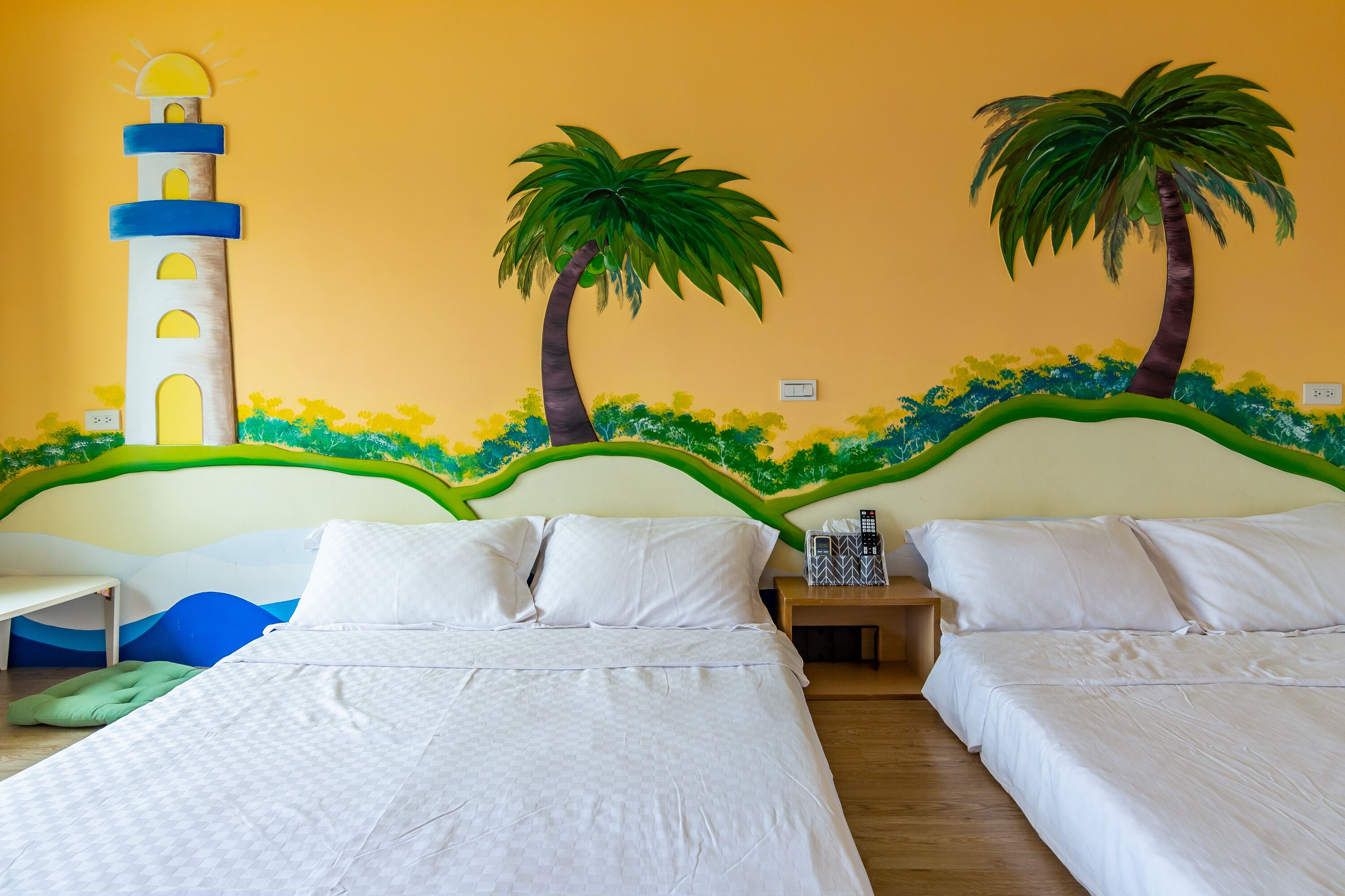 Habitación de hotel con dos camas, una mesita de noche y un mural en la pared con un faro y palmeras.