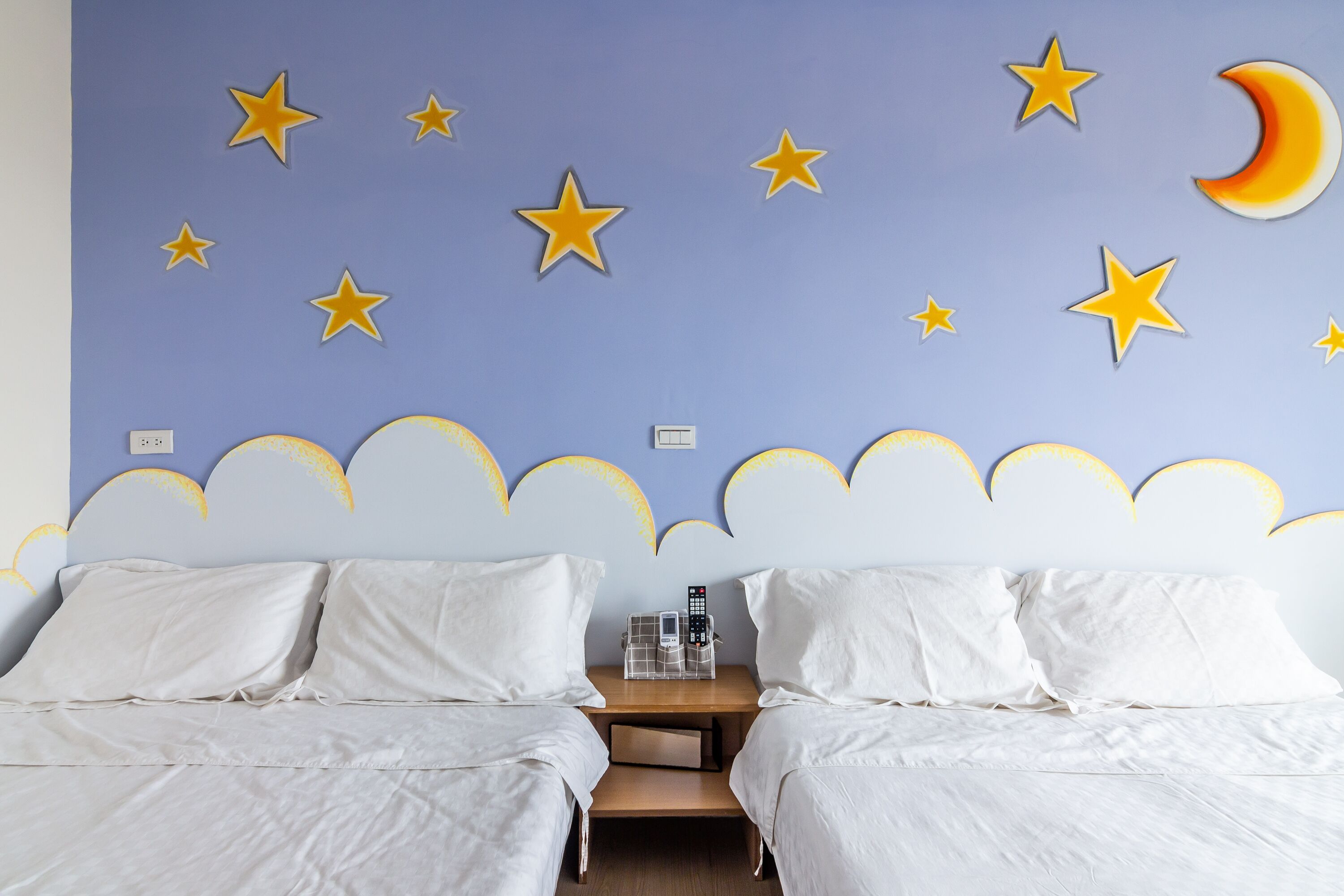 Habitación de hotel con dos camas, una mesita de noche con teléfono y lámpara, y una pared decorada con estrellas y una luna creciente.