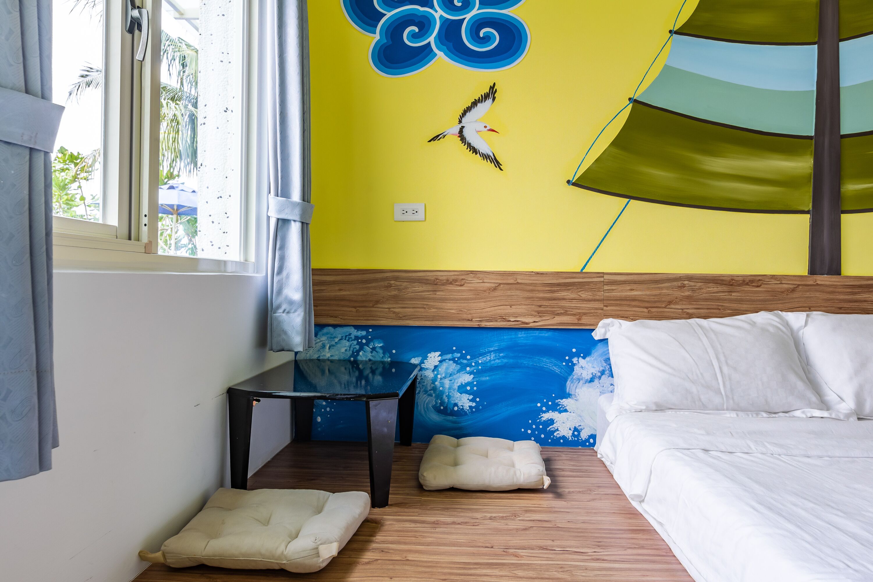 A room with a bed, a small table, two cushions, a window with curtains, and a wall with a blue wave design.