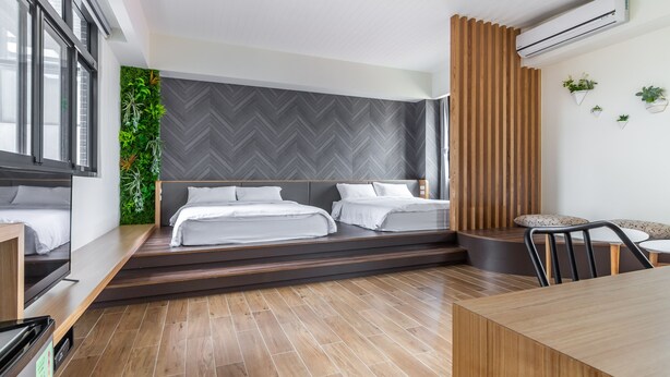 A modern hotel room with two beds, a wooden dining table, a built-in TV, and a green wall with plants.