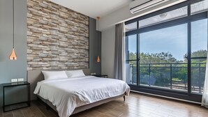 A modern bedroom with a large bed, a wooden wall panel, a nightstand, a bedside table, and a large window with a view of trees.