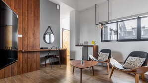 A modern living room with a wooden floor, a round table, two chairs, a TV, and a window with a view of buildings.