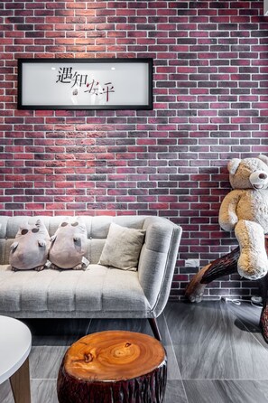 A cozy room with a brick wall, a framed sign, a sofa with cushions, a large teddy bear, and a round wooden table.