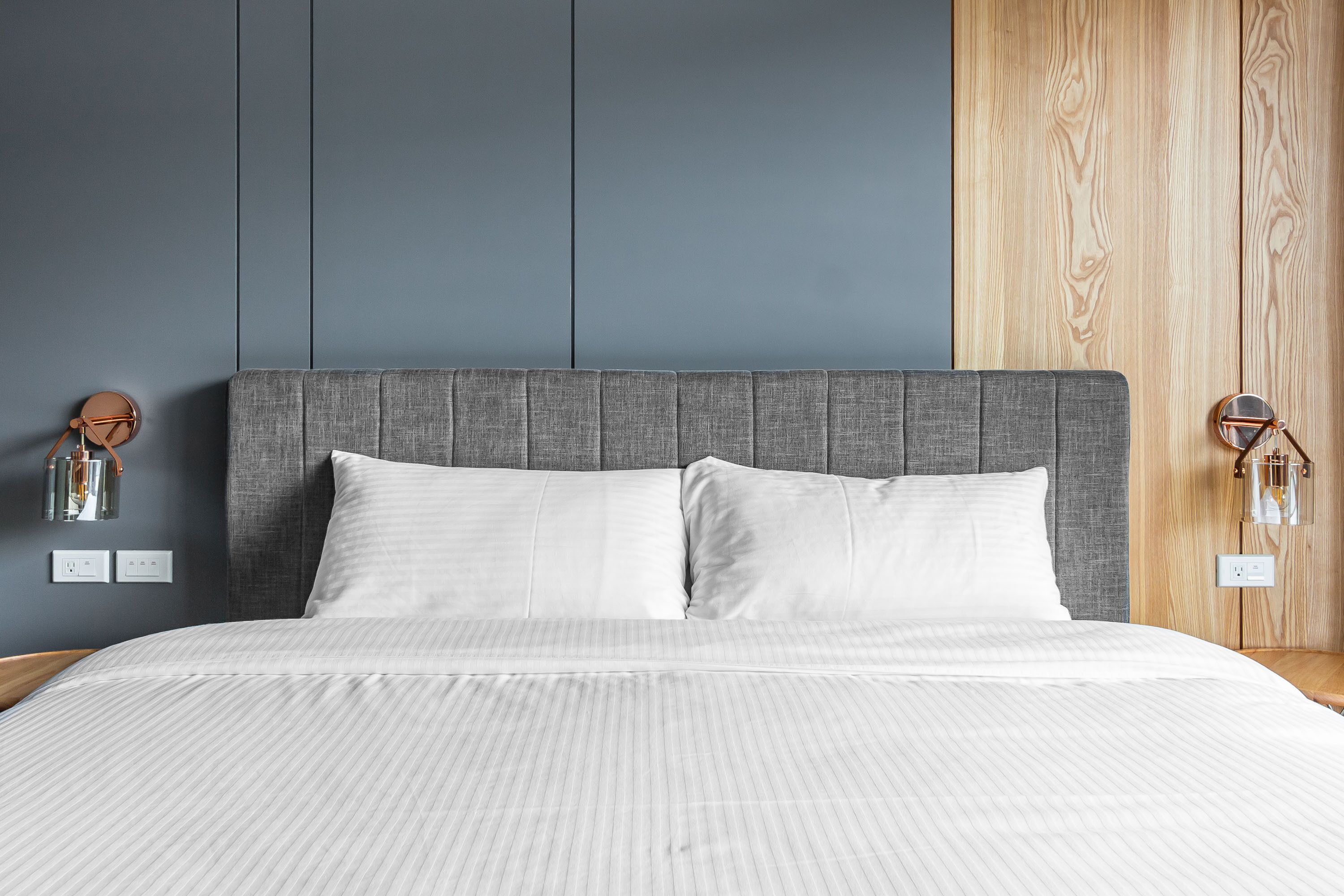 Een net opgemaakt bed met een grijze hoofdbord, wit beddengoed en een houten paneel aan de rechterkant.