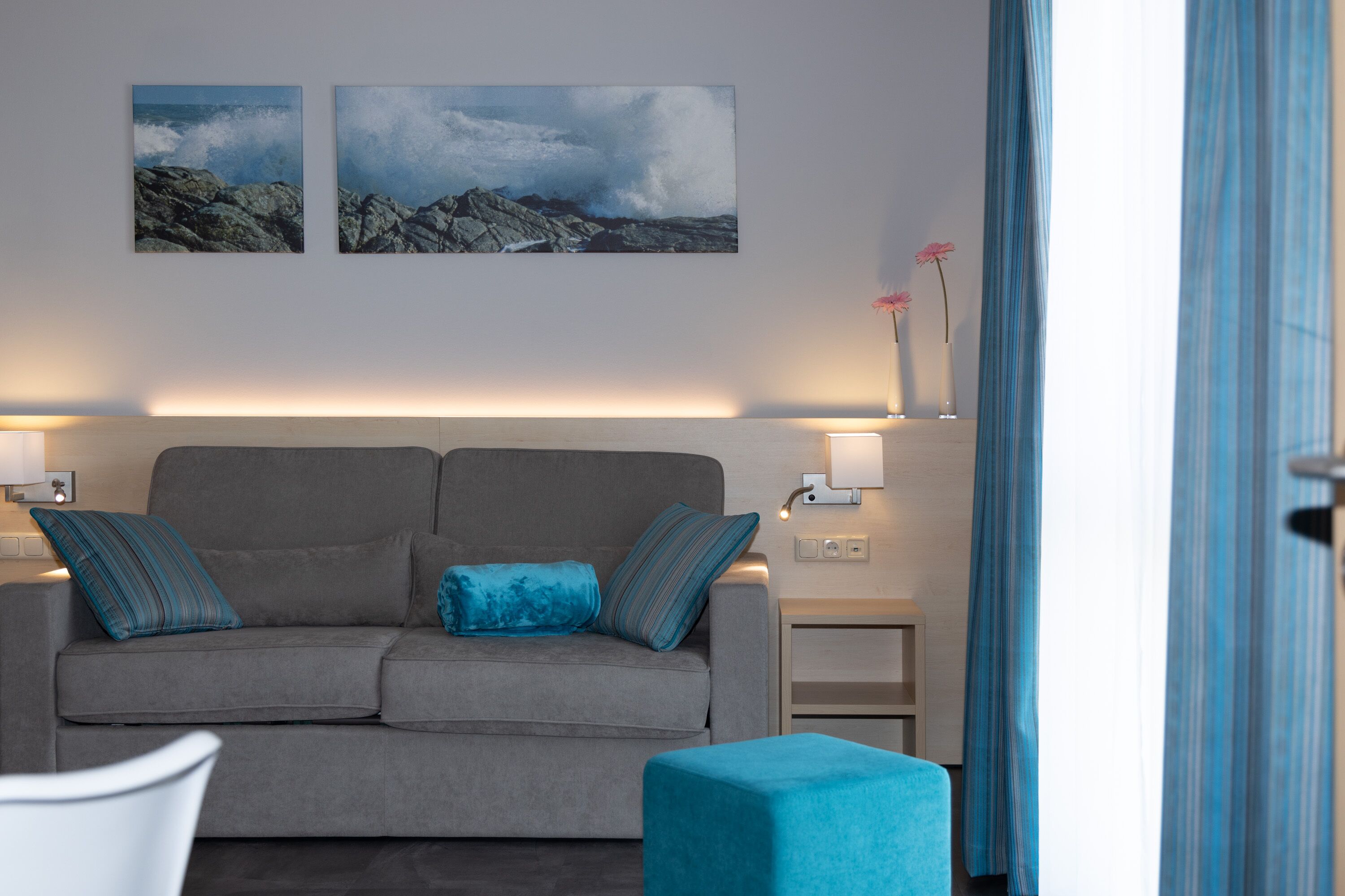 A modern living room with a grey sofa, two blue cushions, a blue ottoman, and two wall-mounted lamps. There are two framed pictures on the wall and a vase with flowers on a side table.