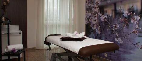 A massage table with towels and a robe, a small table with a vase of flowers, and a window with sheer curtains.