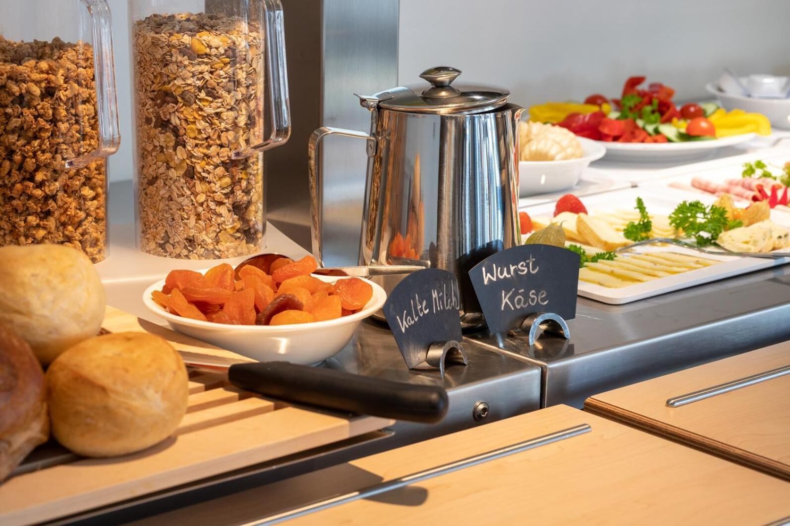 Montaje de bufé con diversas opciones de comida, incluyendo un tazón de albaricoques secos, un recipiente de granola y una olla de sopa.