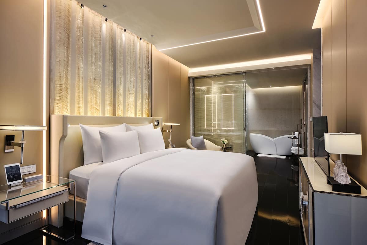 Moderni hotellihuone, jossa on suuri sänky, lasinen työpöytä, lasiseinällinen suihku ja peili.