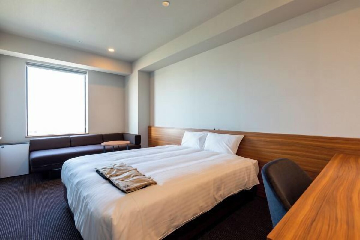 Ein Hotelzimmer mit einem großen Bett, einer kleinen Sitzecke, einem Schreibtisch und einem Fenster, das für natürliches Licht sorgt.