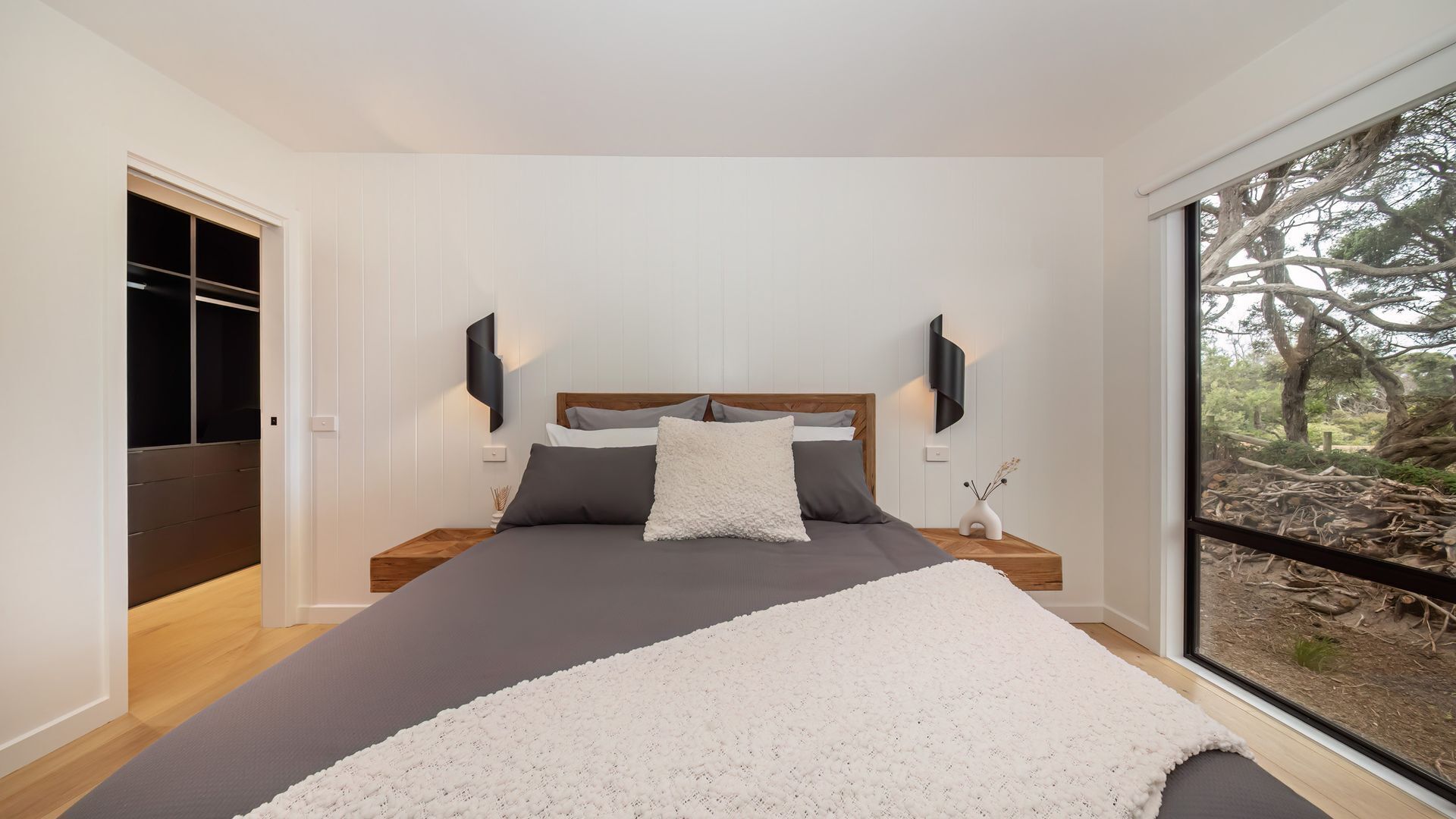 A modern bedroom with a large bed, a wooden nightstand, a wall-mounted TV, and a view of trees through a large window.