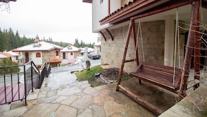 Un espacio al aire libre con suelo de piedra, un conjunto de columpios de madera, una barandilla metálica y vistas a casas vecinas.