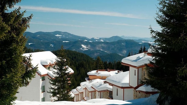 Un resort en la montaña cubierto de nieve con edificios de estilo chalete, vistas despejadas de las montañas circundantes y un paisaje boscoso de fondo.