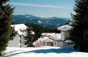 Un resort en la montaña cubierto de nieve con edificios de estilo chalete, vistas despejadas de las montañas circundantes y un paisaje boscoso de fondo.