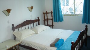 A bedroom with a wooden bed, two bedside tables, a window with blue curtains, and a wall-mounted shelf. - House in Quiet Residence (Grand-Baie)