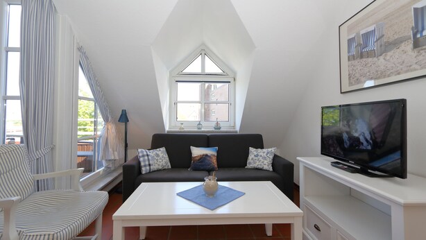 A modern living room with a white ceiling, a large window, a black sofa, a white coffee table, a striped armchair, a TV on a white stand, and a framed picture on the wall. - App. Reif für die Insel (Sylt)