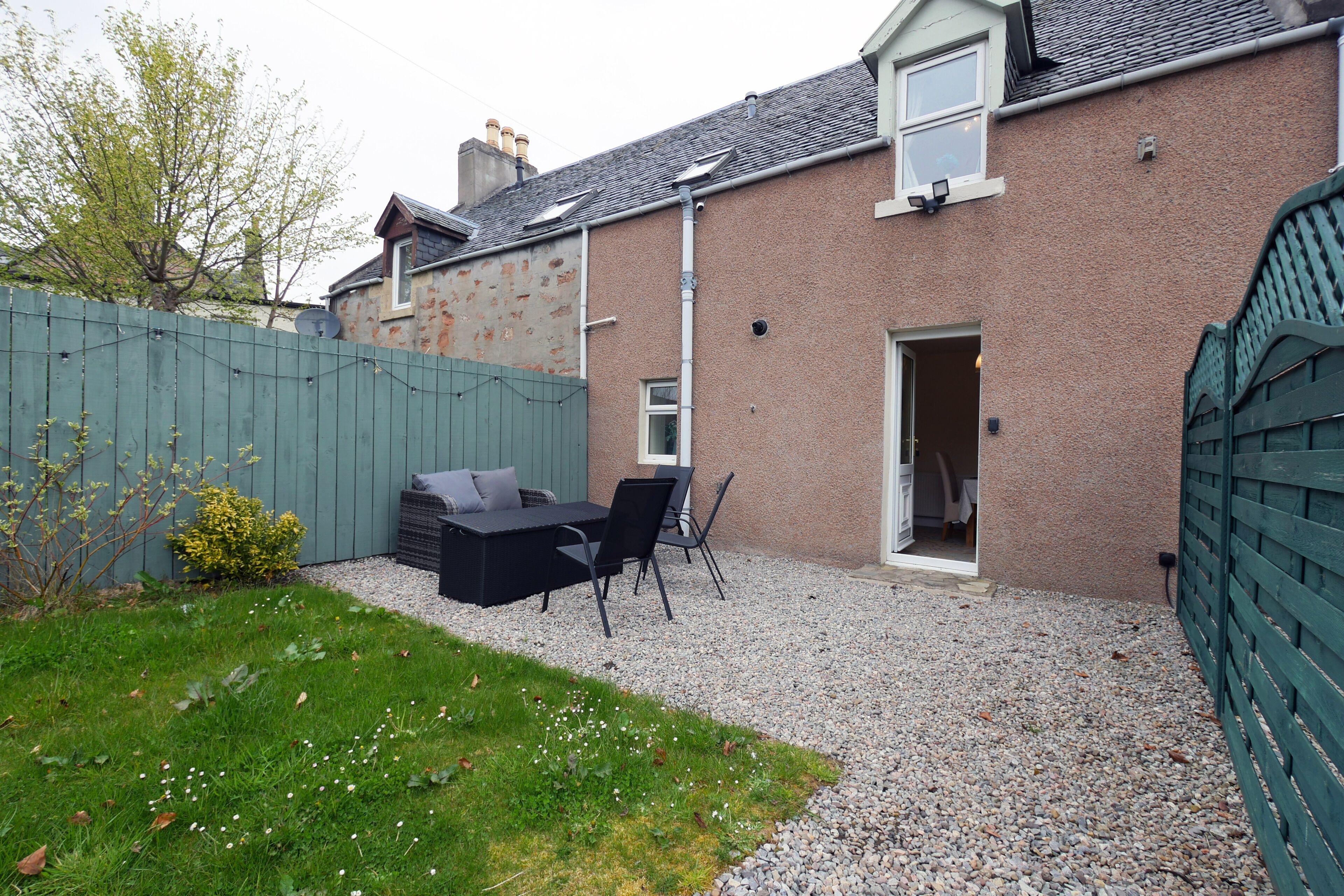 Une maison avec une terrasse en gravier, une clôture verte et un petit jardin.