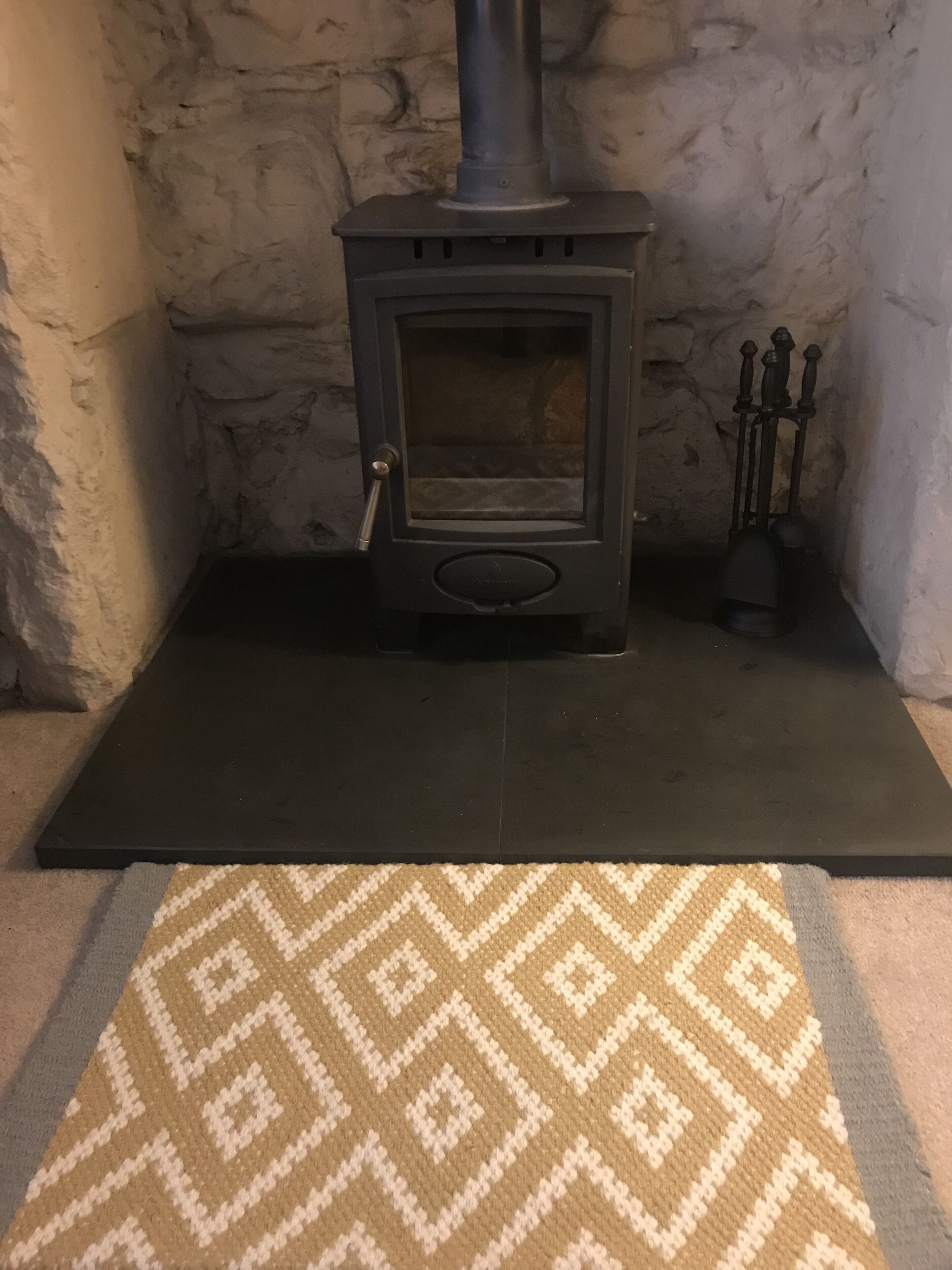 A wood-burning stove with a glass door, set on a black platform against a stone wall.