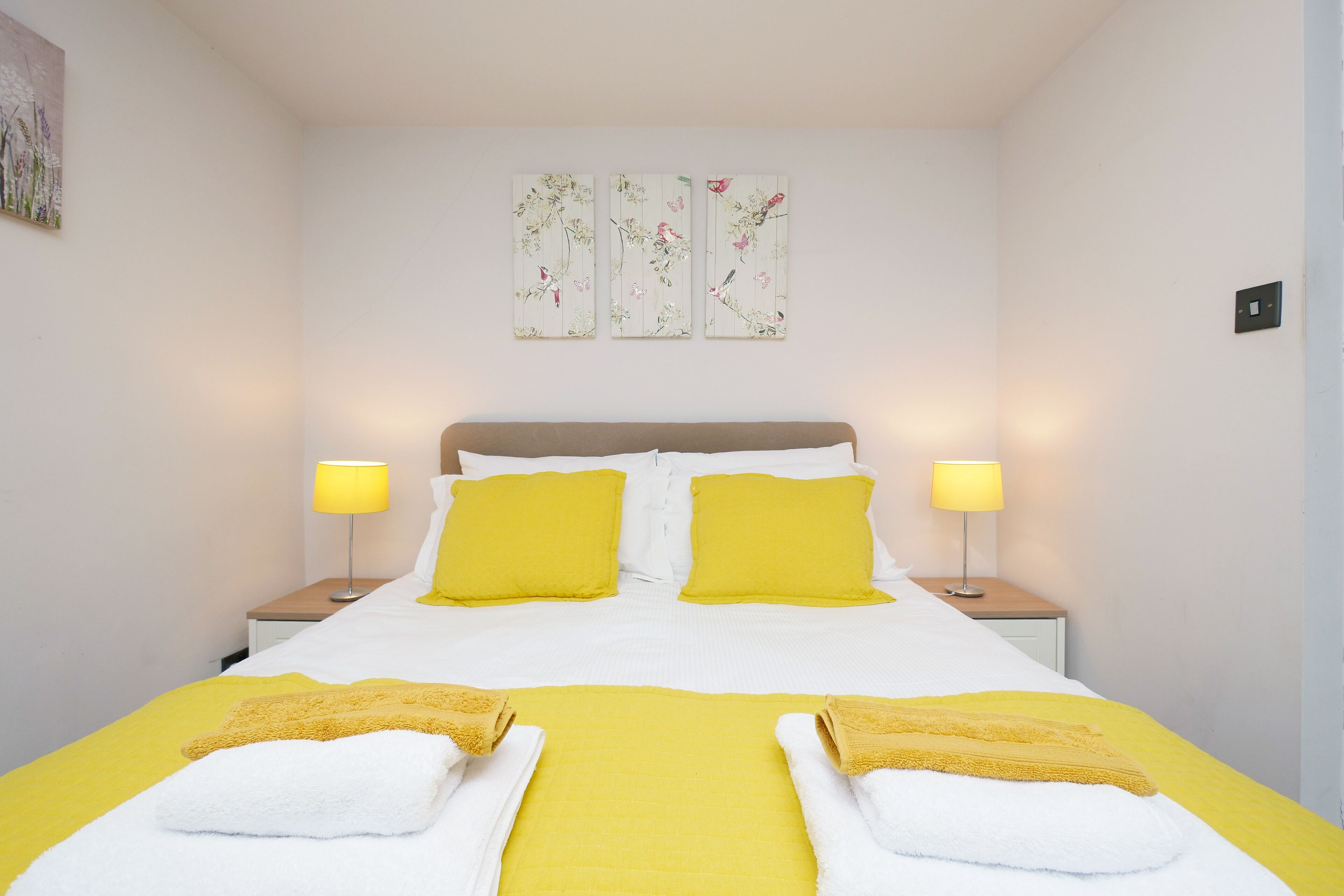 A neatly made bed with yellow and white bedding, two bedside lamps, and three wall-mounted floral artworks.