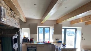 Een ruime keuken met hoge plafonds en een stenen openhaard, een zithoek met een lederen stoel, en grote ramen die zorgen voor veel daglicht.