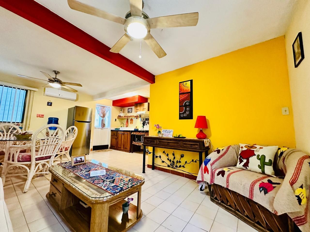 A living room with a ceiling fan, a dining area with chairs, a kitchen in the background, and a sofa with a patterned throw.