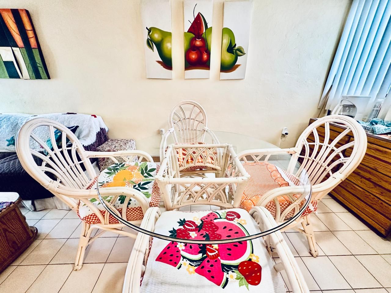 Un área de comedor con sillas de mimbre y una mesa de vidrio, un sofá con una manta estampada y decoración de pared con motivos de frutas.
