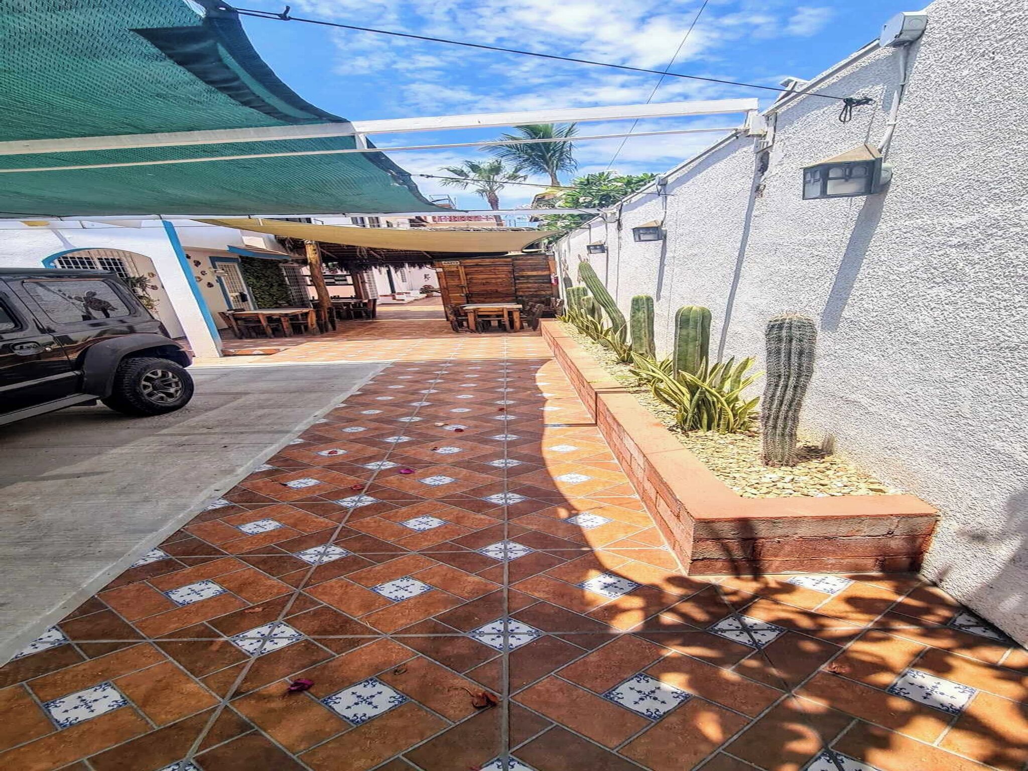 A paved walkway leading to a patio area with outdoor seating, a vehicle parked on the side, and cacti along the wall.