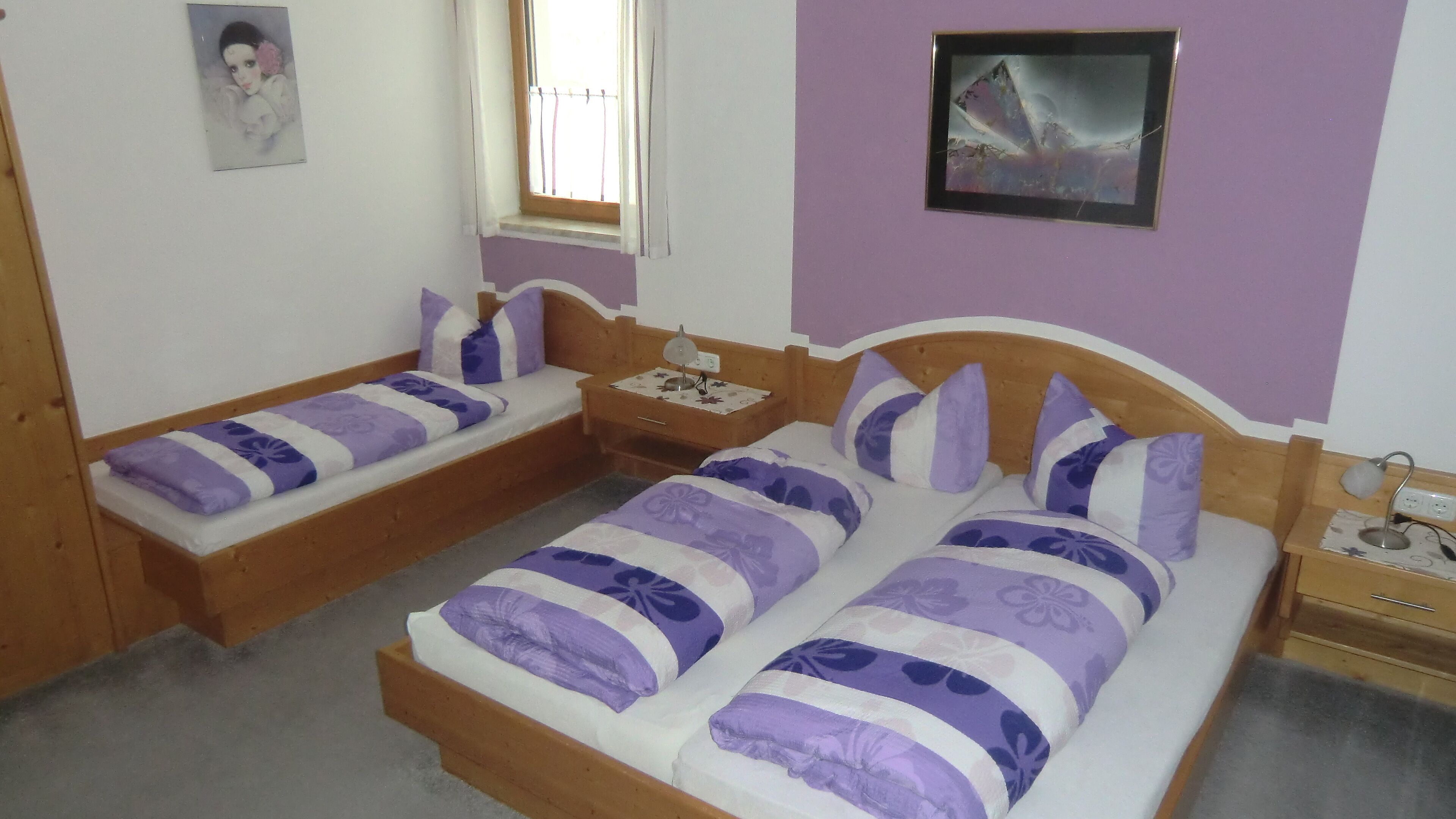 Ein Schlafzimmer mit zwei hölzernen Betten, beide mit violett-weißer Bettwäsche. An einer Seite steht ein kleiner Nachttisch mit einer Lampe, an der anderen ein Beistelltisch. Ein Fenster mit Vorhängen ist sichtbar, und ein gerahmtes Bild hängt an der Wand.