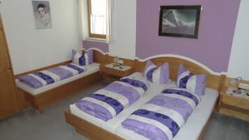 Ein Schlafzimmer mit zwei hölzernen Betten, beide mit violett-weißer Bettwäsche. An einer Seite steht ein kleiner Nachttisch mit einer Lampe, an der anderen ein Beistelltisch. Ein Fenster mit Vorhängen ist sichtbar, und ein gerahmtes Bild hängt an der Wand.