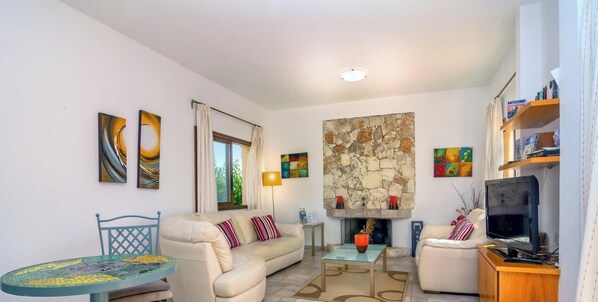 A living room with a sofa, a coffee table, a TV, and a stone fireplace. - Large Pool & Garden, Billiard, Peaceful Area, Near to the Sea, Coral Bay Paphos (Pegeia)