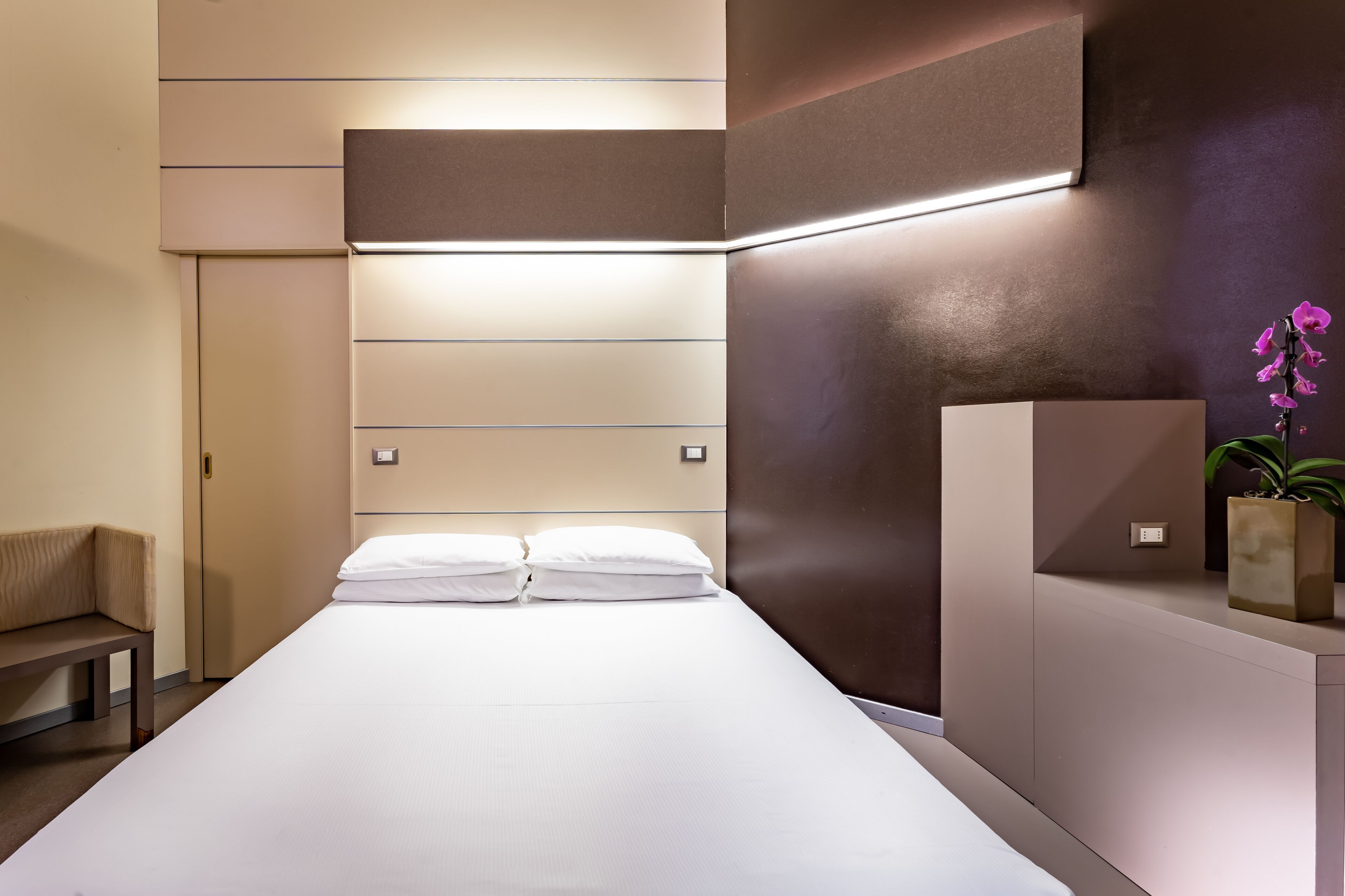 Una habitación de hotel moderna con una cama, mesitas de noche, un banco y una orquídea en maceta.