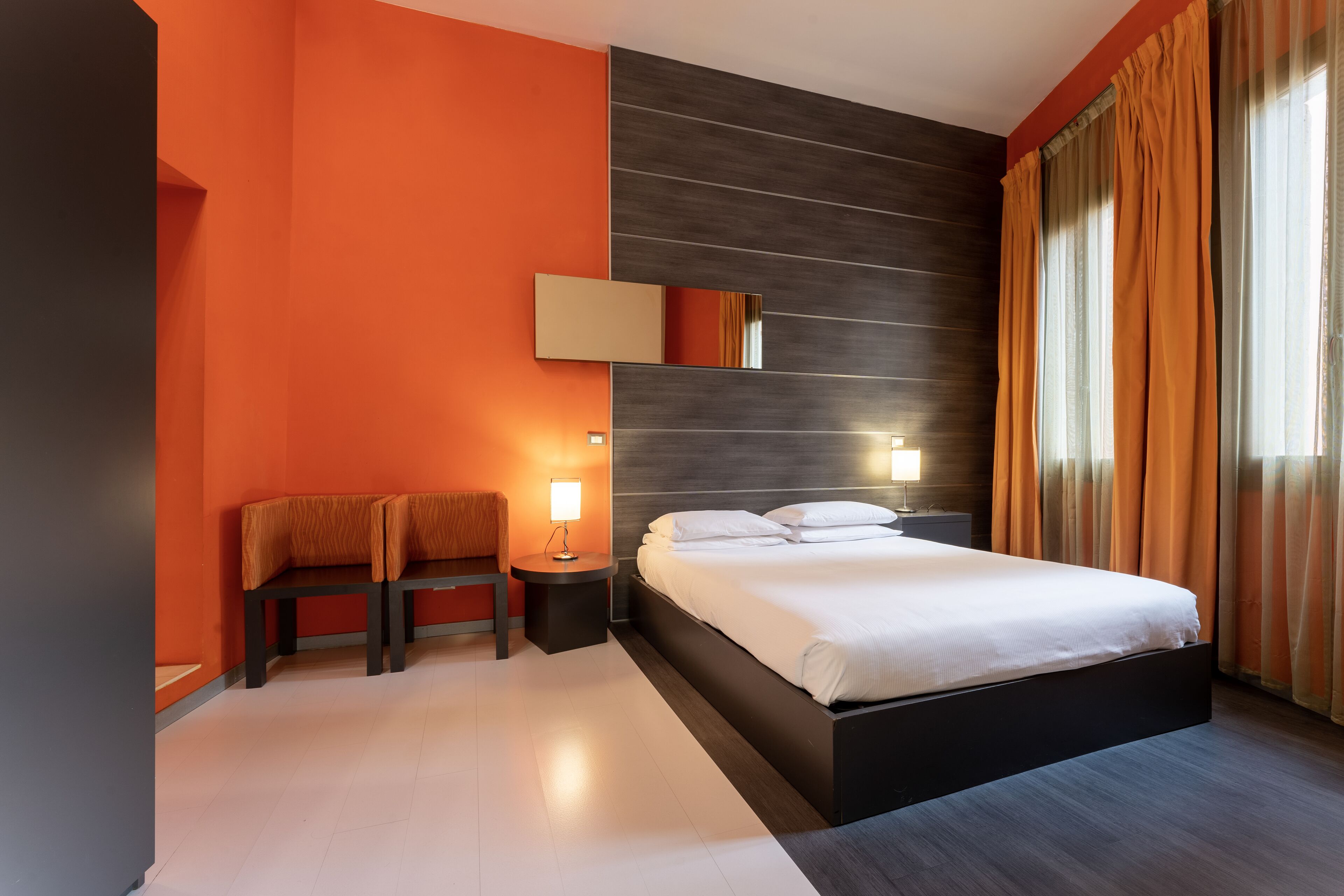 A modern hotel room with a large bed, two bedside tables with lamps, a bench, and a large mirror.