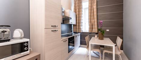 A compact kitchen with a microwave, toaster, and oven. There is a dining table with chairs and a potted plant.