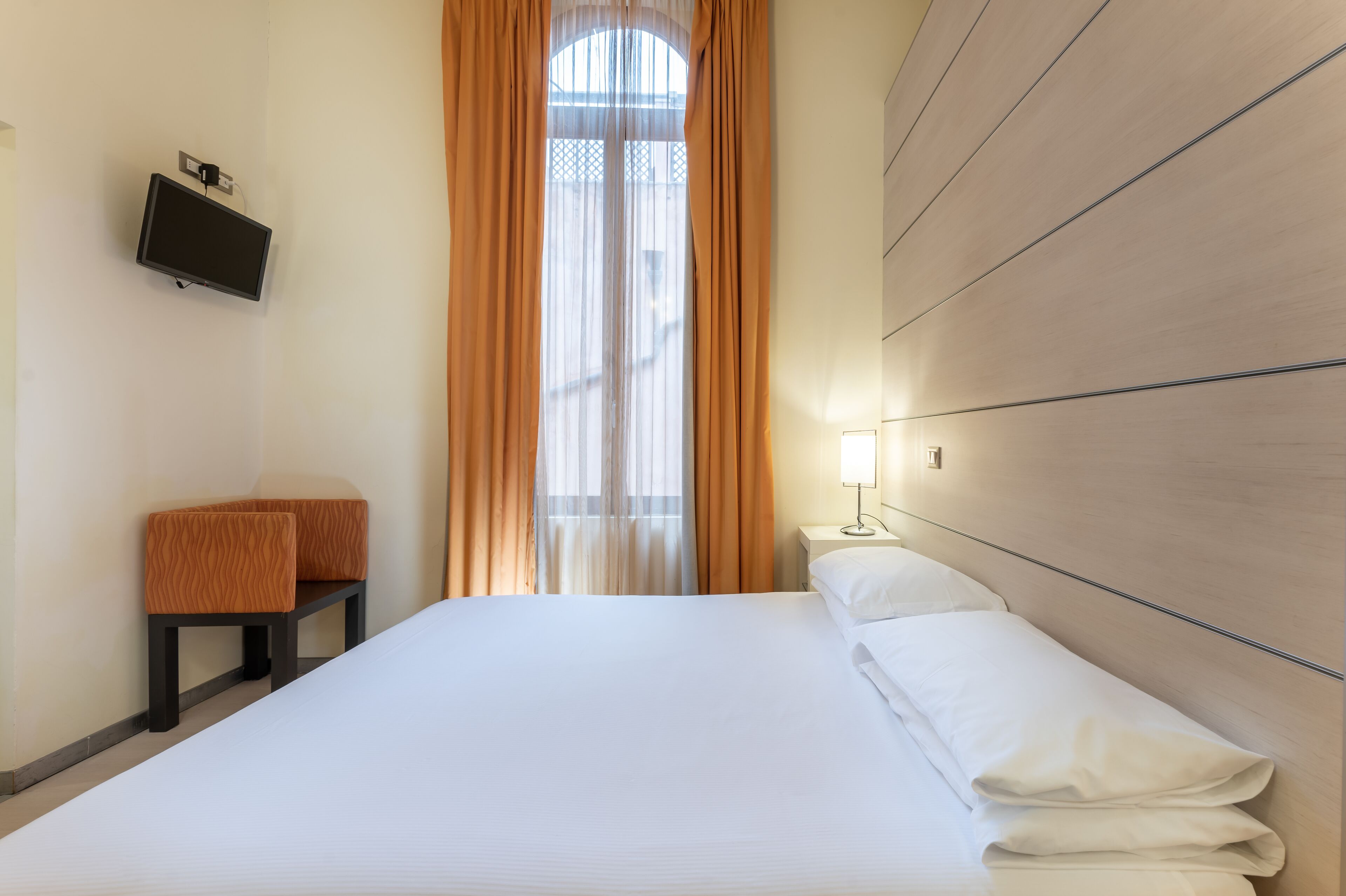 A hotel room with a bed, a TV mounted on the wall, a bedside table with a lamp, and a bench.
