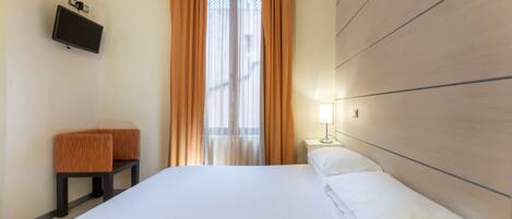 A hotel room with a bed, a TV mounted on the wall, a bedside table with a lamp, and a bench.