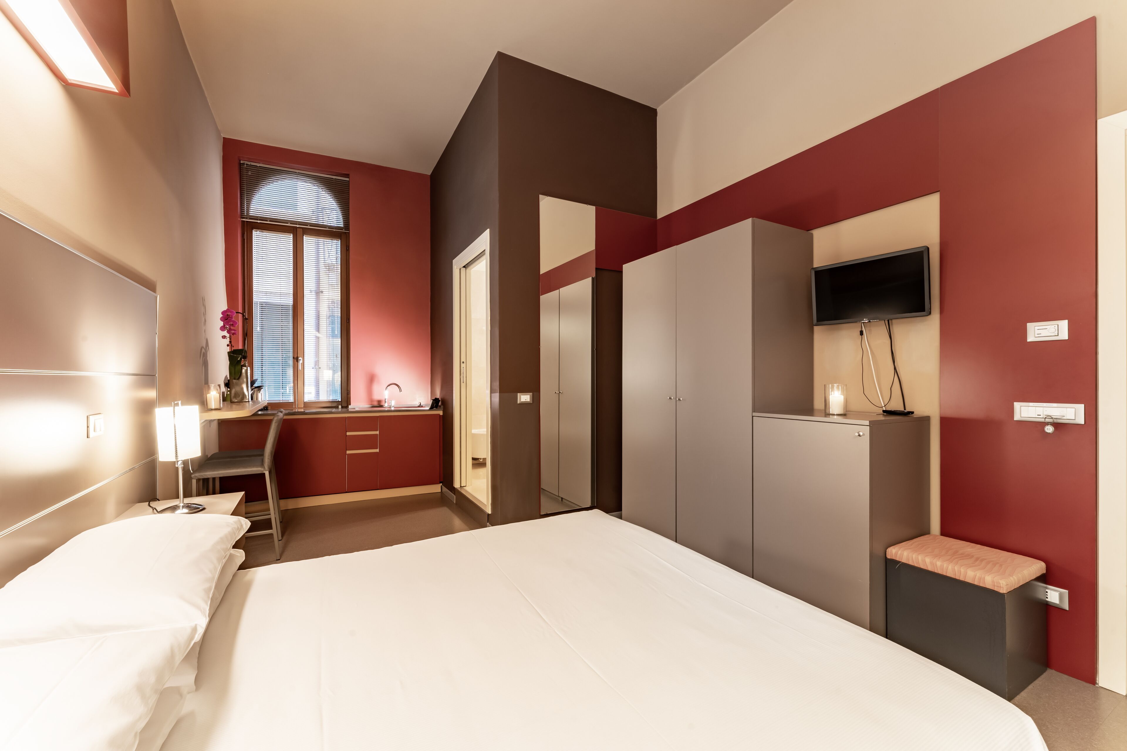 Habitació d'hotel amb un llit, una taula de treball amb cadira, un televisor fixat a la paret i una finestra amb persianes.