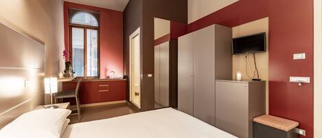 Habitación de hotel con cama, escritorio con silla, televisor montado en la pared y ventana con persianas.