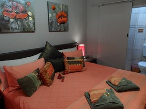 Una cama bien tendida con varios almohadones y una colcha, una lámpara de noche y dos cuadros en la pared.