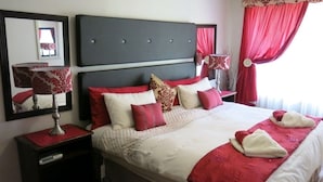 Et hotelværelse med en stor seng, rødt og hvidt sengetøj, et natbord med lampe og et spejl på væggen.